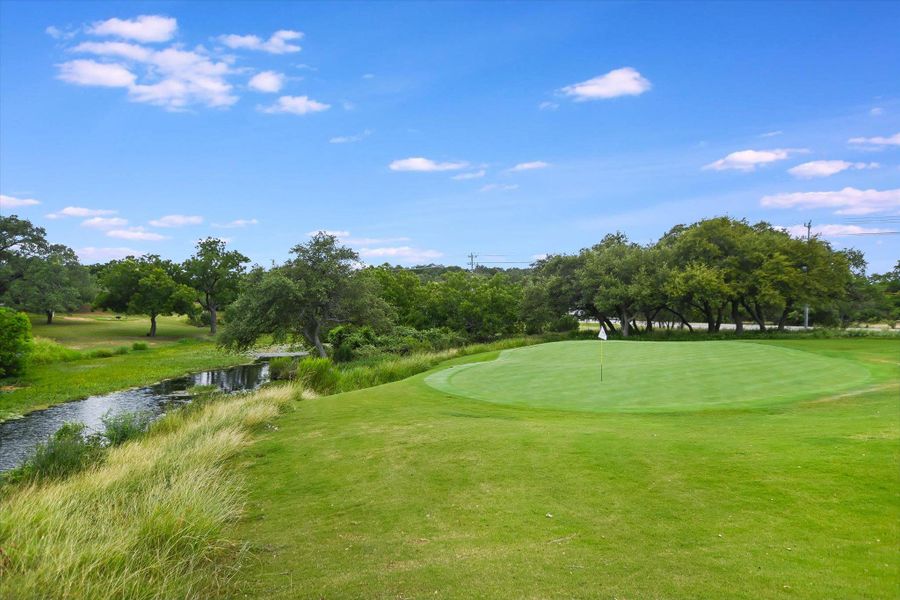 A pristine golf course green borders a tranquil stream, framed by verdant trees and a clear sky.
