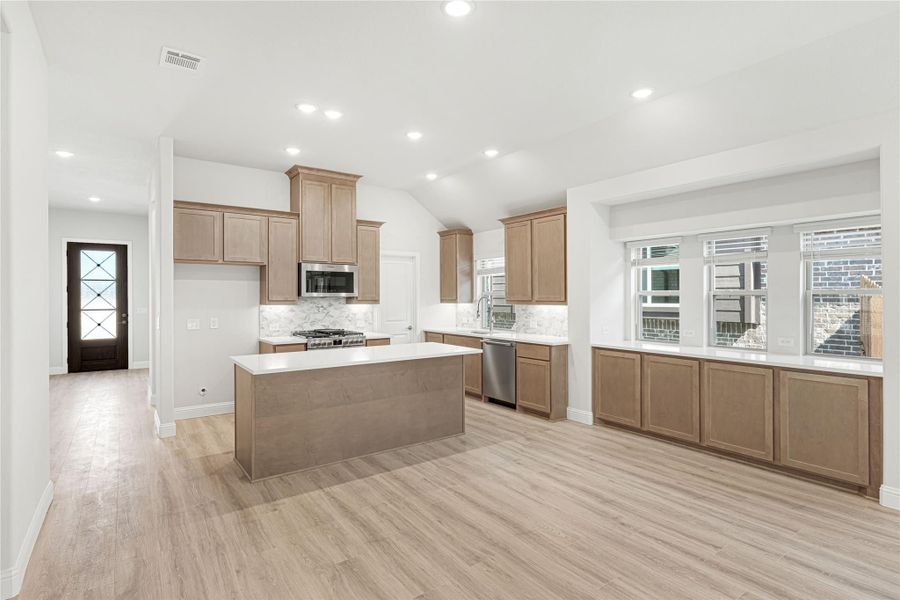 Open kitchen with tan wood cabinets, center island, stainless appliances, and light hardwood floors Open kitchen with tan wood cabinets, center island, stainless appliances, and light hardwood floors