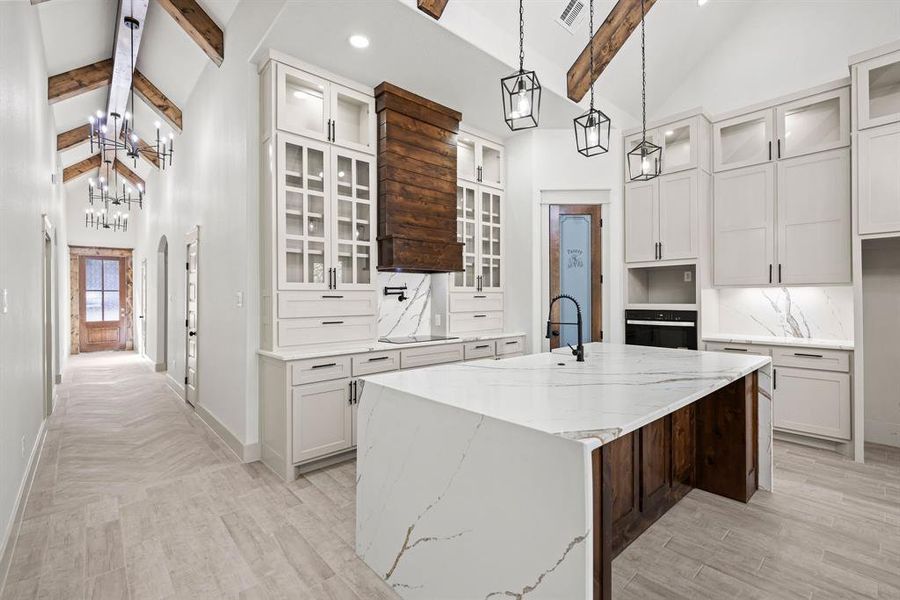 Kitchen with high vaulted ceiling, beamed ceiling, light stone countertops, an island with sink, and glass insert cabinets