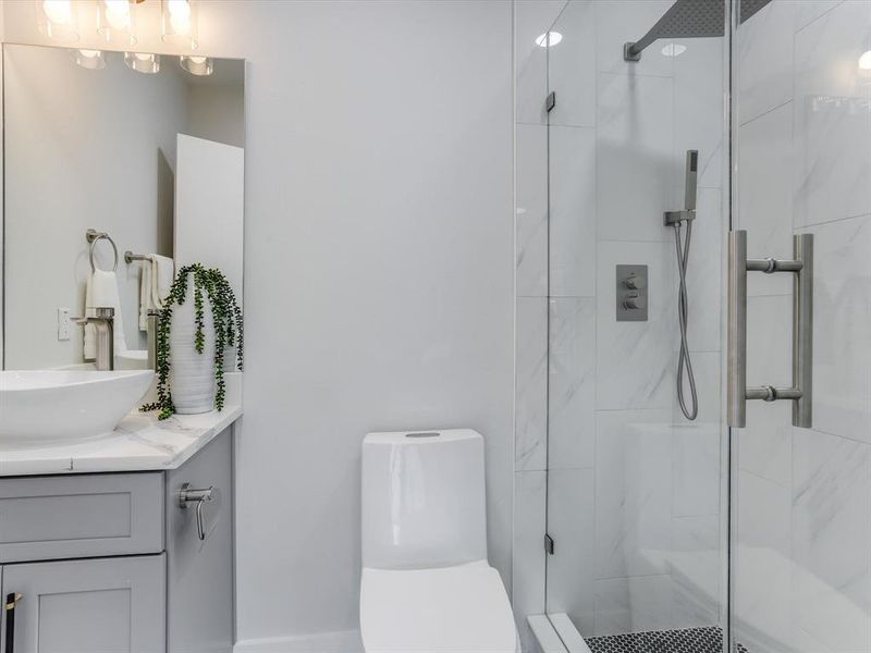Contemporary bathroom featuring a frameless glass shower enclosure with a rainfall showerhead and handheld sprayer, a white vessel sink on a stone countertop, and gray shaker-style vanity cabinetry Contemporary bathroom featuring a frameless glass shower enclosure with a rainfall showerhead and handheld sprayer, a white vessel sink on a stone countertop, and gray shaker-style vanity cabinetry