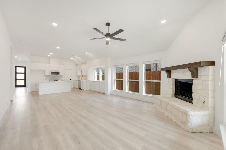 Open living area with stone fireplace, ceiling fan, light wood floors, and open kitchen in background Open living area with stone fireplace, ceiling fan, light wood floors, and open kitchen in background