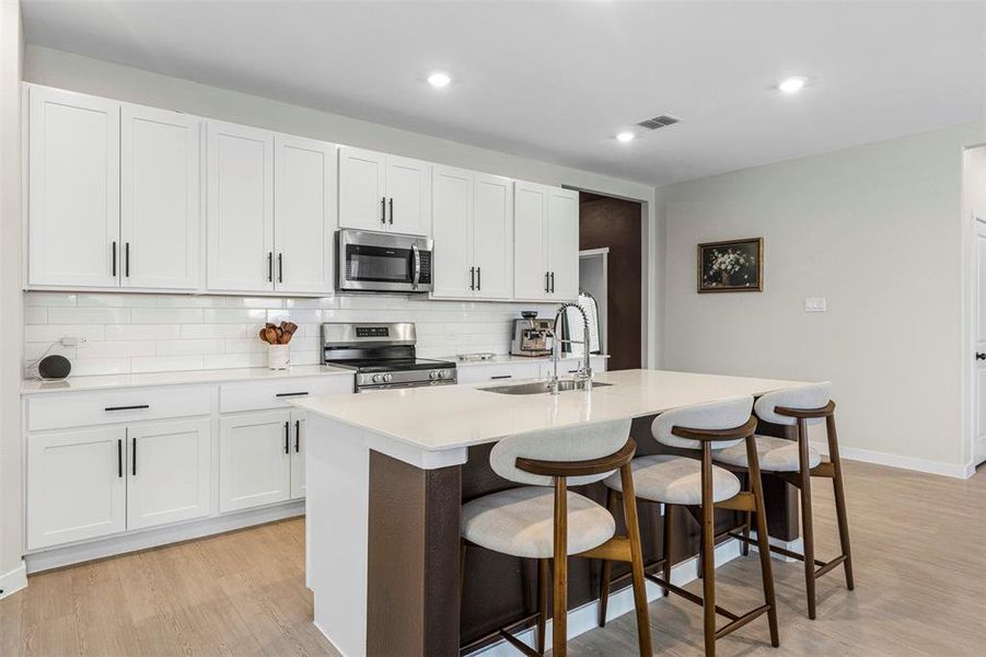 Modern kitchen featuring an expansive island with seating, white shaker cabinetry, stainless steel appliances, a subway tile backsplash, and light wood-finish flooring Modern kitchen featuring an expansive island with seating, white shaker cabinetry, stainless steel appliances, a subway tile backsplash, and light wood-finish flooring