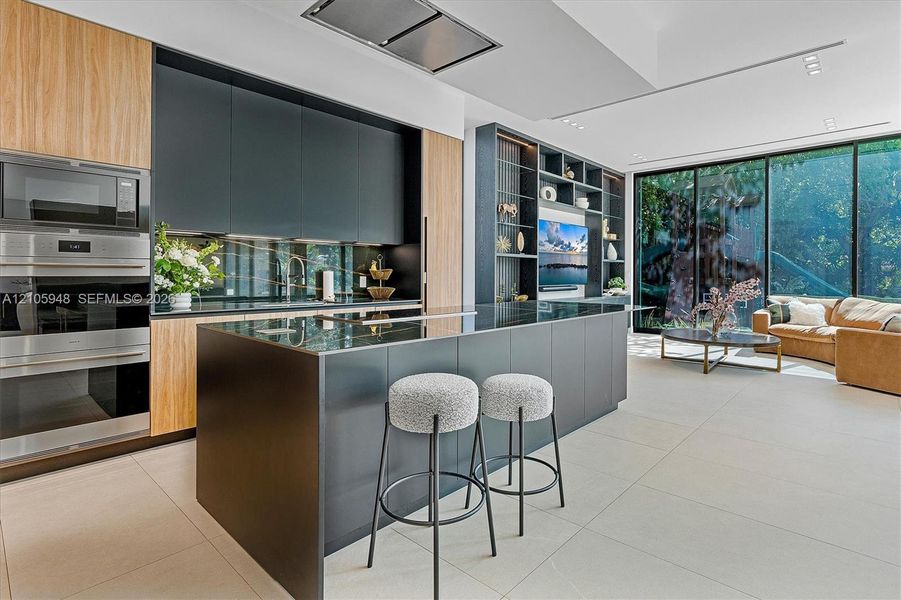 Contemporary kitchen featuring matte cabinetry, wood-finish paneling, a large island with a surface-mounted cooktop, floor-to-ceiling glass sliding doors, and custom floor-to-ceiling built-in shelving