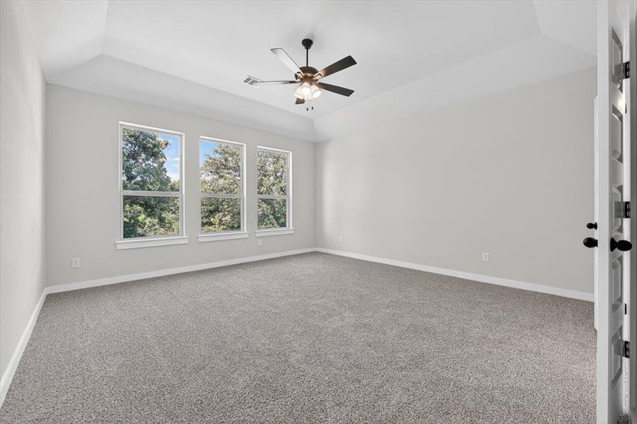 Spacious room featuring a tray ceiling, three large windows, textured carpet flooring, and a ceiling fan with light fixture