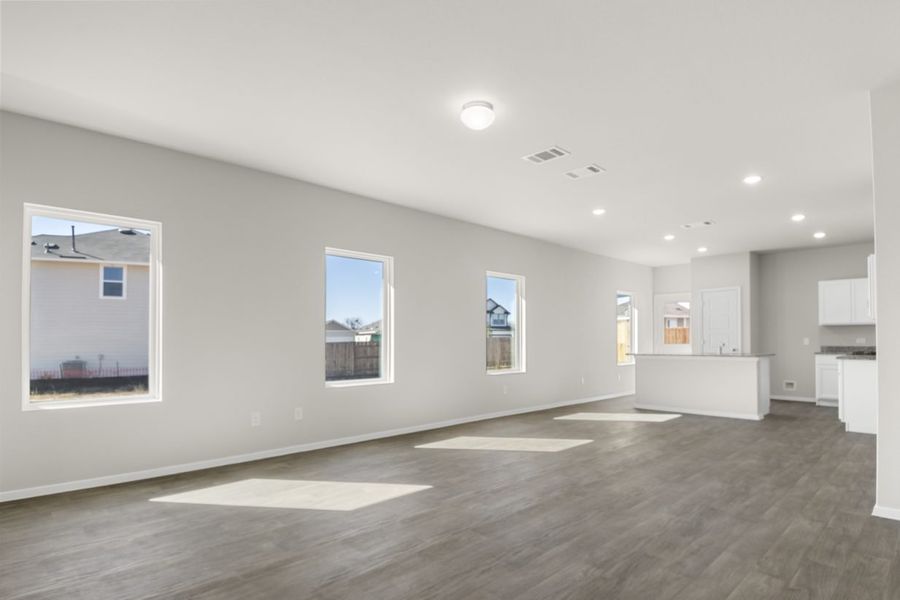 Image of a living room with cream walls, dark brown flooring, white trim, windows and a kitchen in the distance