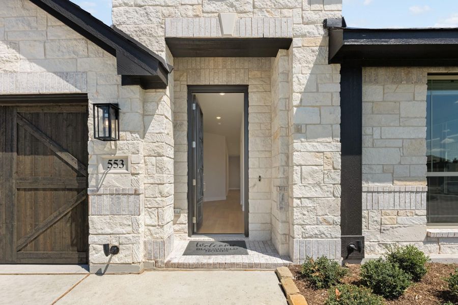Front entry of stone home with gray door, welcome mat, wall lantern, and wood garage door beside it.