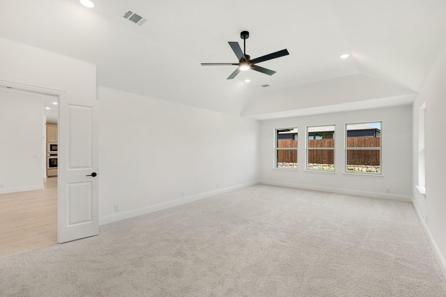 Spacious bedroom with vaulted ceiling, ceiling fan, carpet flooring, and three windows overlooking backyard fence.