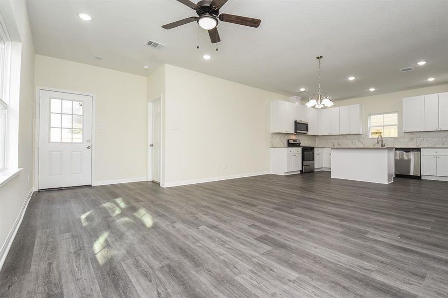 A spacious empty room with gray flooring, white kitchen cabinets, modern appliances, and a ceiling fan, illuminated by natural light.