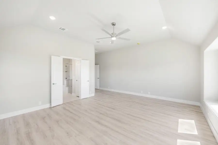 Empty bedroom with vaulted ceiling, ceiling fan, light wood floors, and open ensuite bathroom door