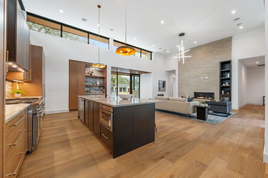 Kitchen with light stone counters, decorative light fixtures, stainless steel appliances, a stone fireplace, and a kitchen island with sink