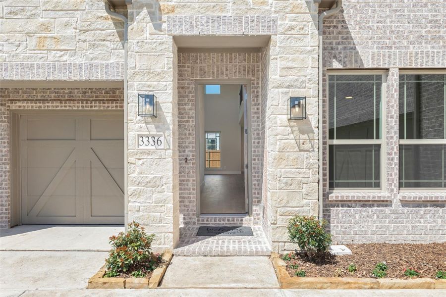 Front entry of a light brick home with open door, house number 3336, wall sconces, and attached garage Front entry of a light brick home with open door, house number 3336, wall sconces, and attached garage