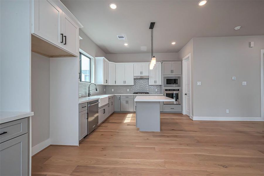 Discover this modern, open-concept kitchen and living area featuring light wood flooring, sleek white cabinetry, a central island with elegant pendant lighting, and stainless steel appliances. A stylish staircase enhances the room's spacious feel.