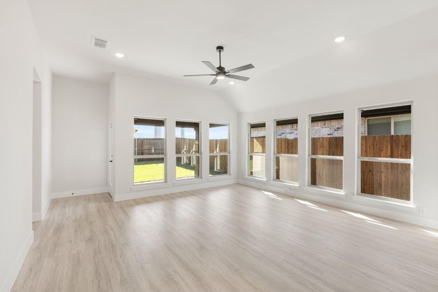 Empty living room with vaulted ceiling, ceiling fan, light wood floors, and large windows overlooking backyard