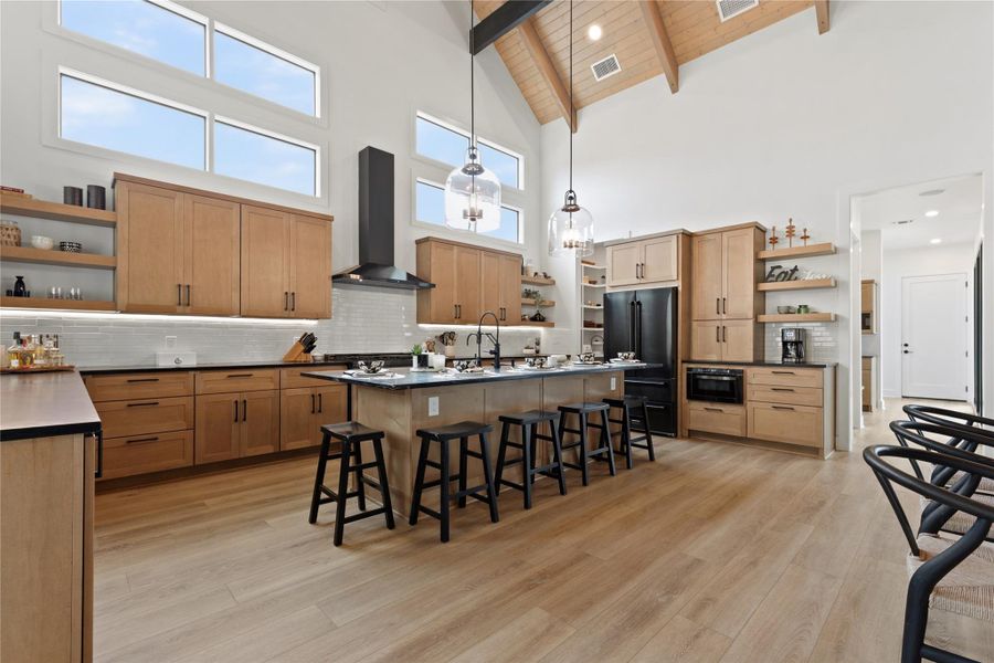 Kitchen featuring a vaulted ceiling with exposed wood beams, high transom windows, light wood-finish flooring, a large island with seating, and integrated appliances Kitchen featuring a vaulted ceiling with exposed wood beams, high transom windows, light wood-finish flooring, a large island with seating, and integrated appliances