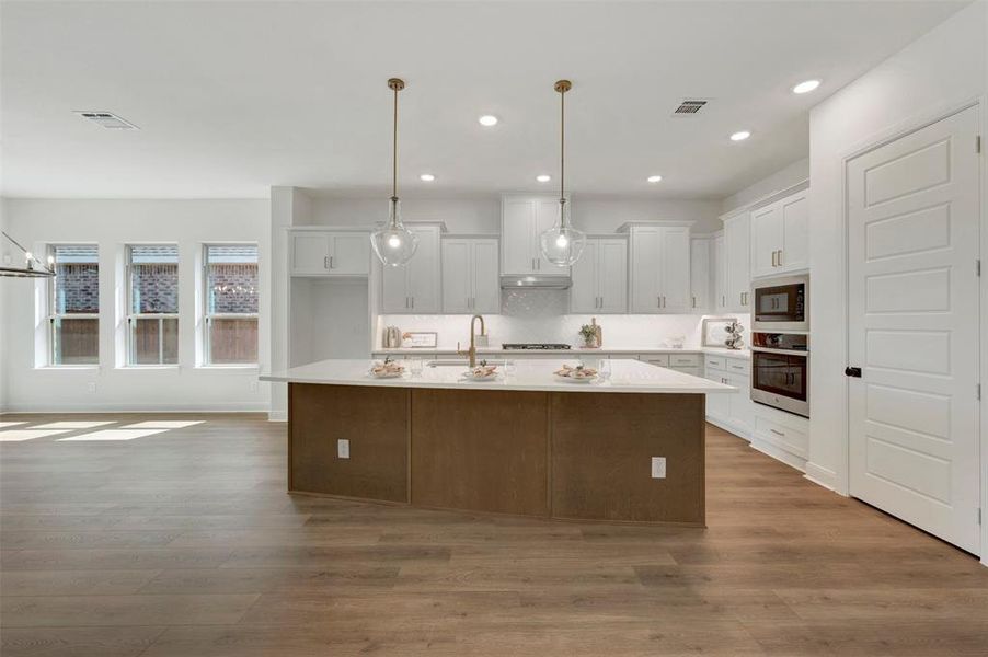 Open-concept kitchen featuring a large island with a light-toned countertop, white cabinetry, integrated stainless steel appliances, and wood-finish flooring Open-concept kitchen featuring a large island with a light-toned countertop, white cabinetry, integrated stainless steel appliances, and wood-finish flooring