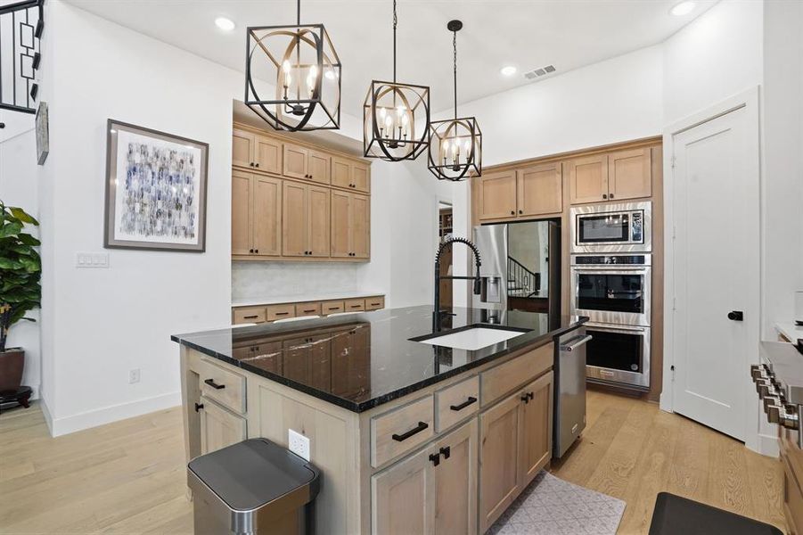 Kitchen with dark stone counters, light brown cabinets, light wood-style flooring, an island with sink, and decorative light fixtures Kitchen with dark stone counters, light brown cabinets, light wood-style flooring, an island with sink, and decorative light fixtures