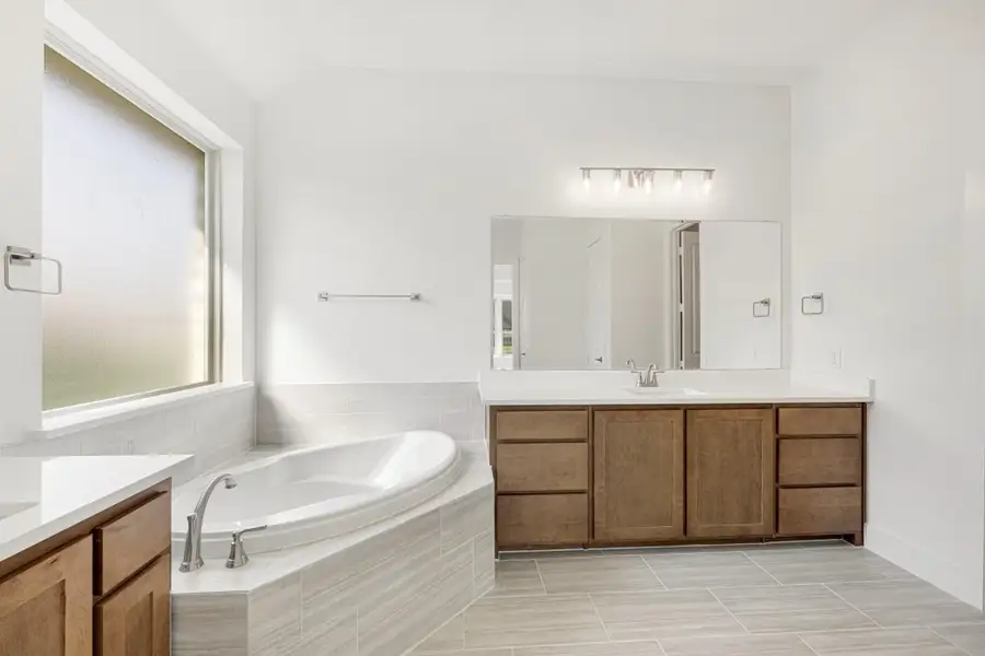 Master bathroom with corner soaking tub, wood vanity, large mirror, and light tile floors Master bathroom with corner soaking tub, wood vanity, large mirror, and light tile floors