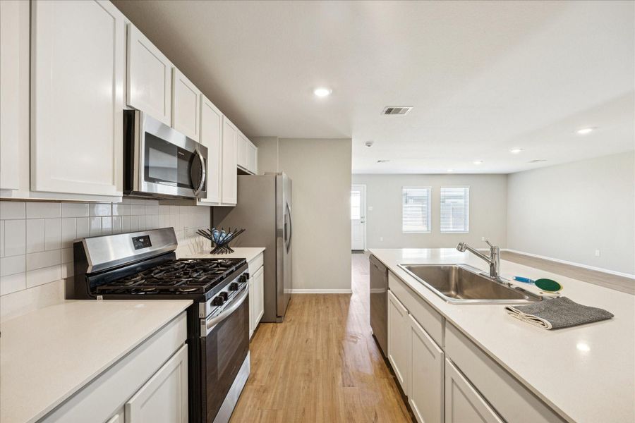 This modern kitchen features sleek stainless steel appliances, white cabinetry, and a spacious open layout with a central island and sink, flowing seamlessly into a bright living area with ample natural light.