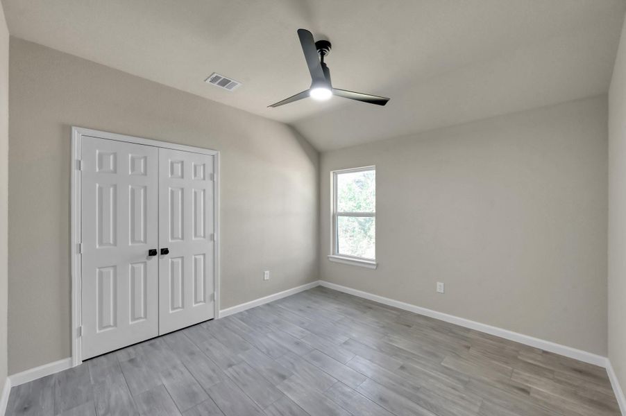 A bright and airy bedroom featuring a vaulted ceiling and a sleek, three-blade modern ceiling fan. Large windows invite plenty of natural light, perfectly complementing the cool-toned flooring and neutral palette.
