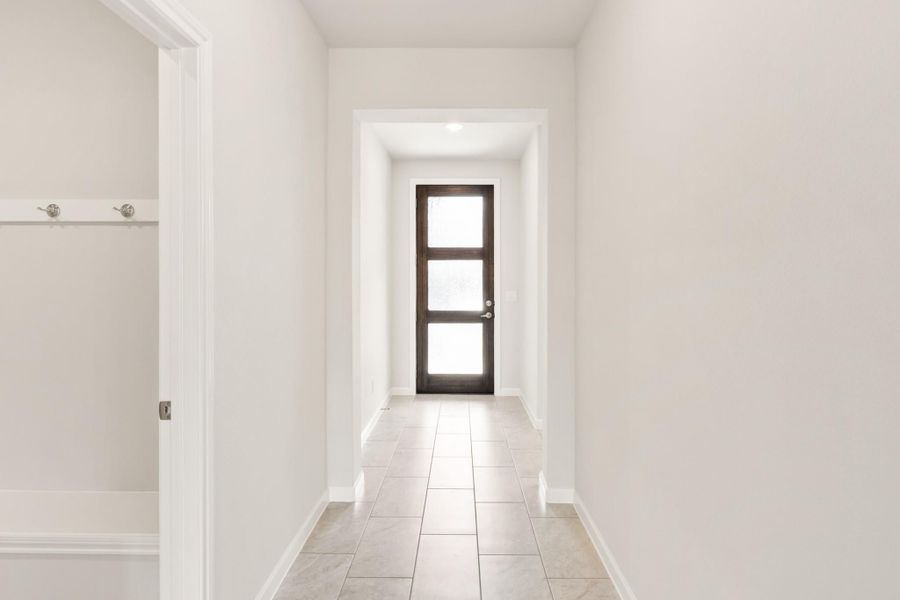 Narrow hallway with tile floors, white walls, and dark wood glass-panel front door at the end Narrow hallway with tile floors, white walls, and dark wood glass-panel front door at the end
