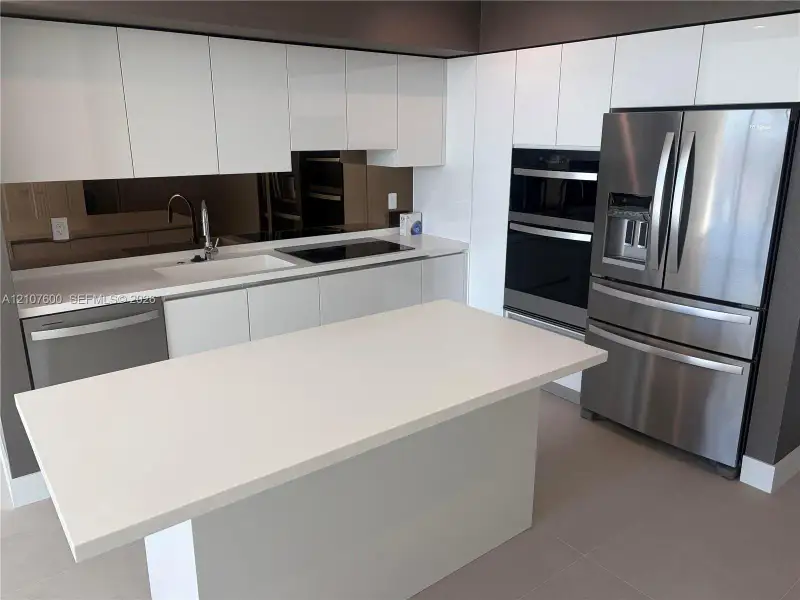 Kitchen featuring white high-gloss cabinetry, stone-surface countertops, a mirrored backsplash, stainless steel appliances, and a central island