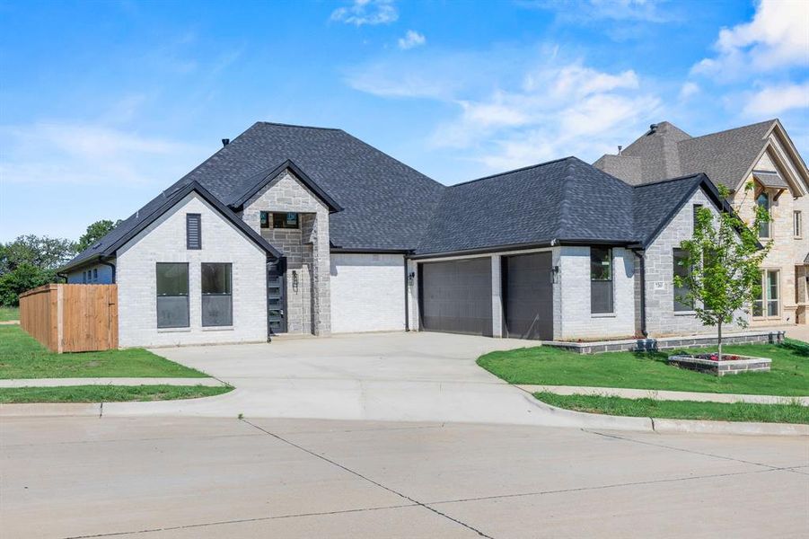 Contemporary residence featuring light-colored brick and stone veneer, a dark shingle roof, a two-car garage with dark doors, a wooden privacy fence, and a concrete driveway Contemporary residence featuring light-colored brick and stone veneer, a dark shingle roof, a two-car garage with dark doors, a wooden privacy fence, and a concrete driveway
