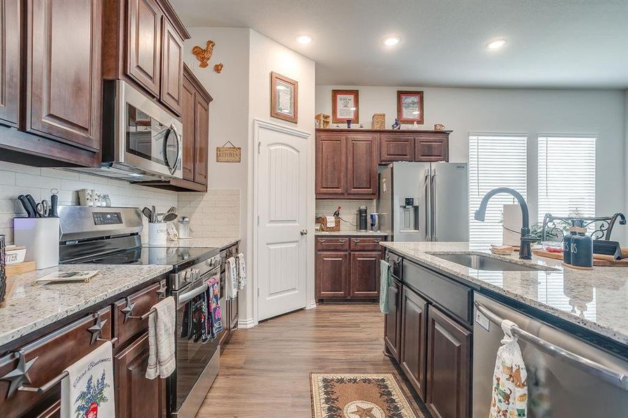 Kitchen with stainless steel appliances, dark brown cabinets, backsplash, light stone countertops, and light wood-type flooring Kitchen with stainless steel appliances, dark brown cabinets, backsplash, light stone countertops, and light wood-type flooring