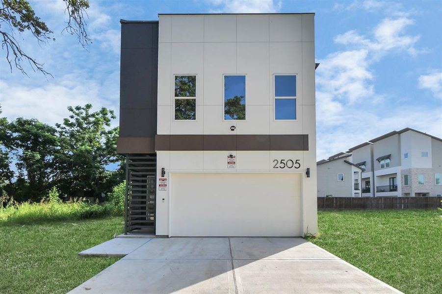 This is a modern two-story residential property with a unique architectural design, featuring a flat roof and a combination of dark and light exterior finishes. The home includes a driveway and is situated on a street with grassy areas and other residential properties in view. Large trees provide a touch of greenery to the neighborhood.