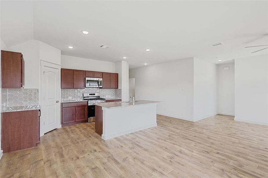 Kitchen featuring decorative backsplash, light wood-type flooring, light stone counters, stainless steel appliances, and a center island with sink