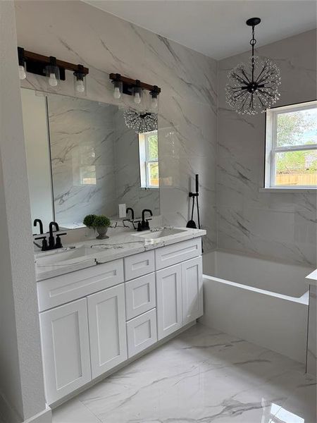 This is a modern bathroom featuring marble-style tiling, a double vanity with white cabinetry, black fixtures, and a stylish chandelier. A large mirror spans the width of the vanity, enhancing the space's elegance and brightness.