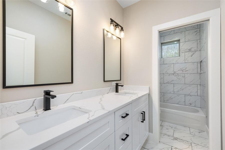 The modern secondary bathroom boasts stylish dual-sinks with sleek black fixtures and large mirrors that enhance the space. A tub-shower combo, adorned with elegant marble-inspired tiles, adds a touch of luxury. Ample natural light floods the room, highlighting the clean, contemporary design and creating a bright, inviting atmosphere.