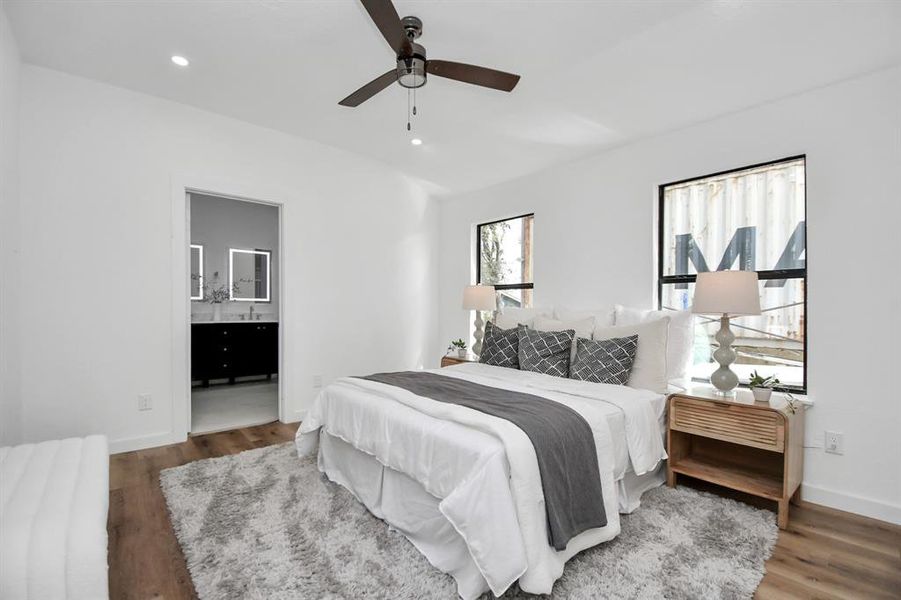 This is a modern, minimalist bedroom featuring clean lines, a neutral color palette with a large abstract wall art piece, and hardwood floors. It includes a ceiling fan and has a view into another room, suggesting a connected floor plan.