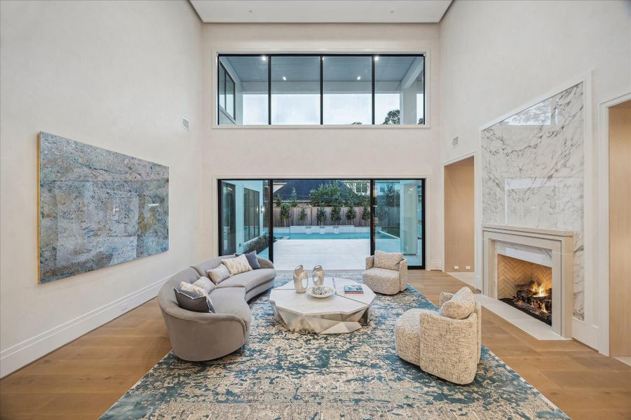 The spacious formal living room showcases soaring plaster ceilings with LED details, plaster walls, expansive windows, and a marble-surround fireplace. Sliding doors open to the patio and pool, creating a seamless indoor–outdoor flow.