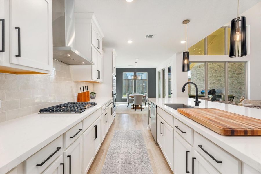 This kitchen features a sleek, modern design with white cabinetry and countertops. It includes a gas cooktop, stylish pendant lighting, and a spacious island with a built-in sink. Large windows provide natural light, and there's an open view into a dining area, creating an inviting and functional space for cooking and entertaining.