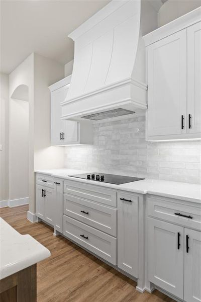 Kitchen with custom range hood, white cabinetry, light wood finished floors, decorative backsplash, and black electric cooktop