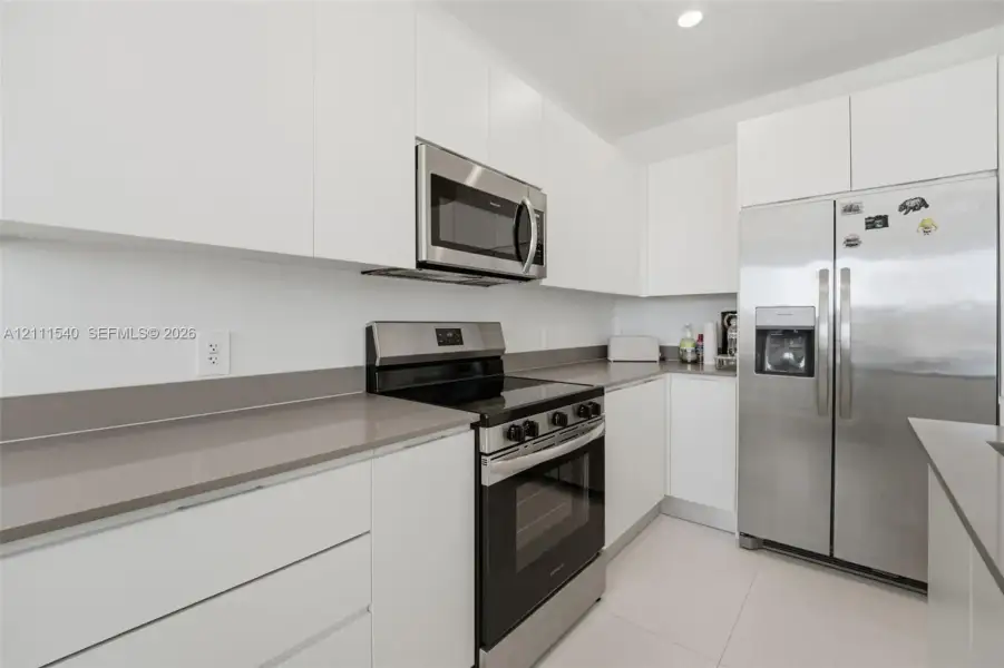 Kitchen featuring flat-panel white cabinetry, solid-surface countertops, recessed lighting, and stainless steel appliances including a range, microwave, and refrigerator