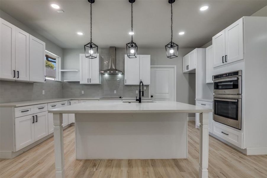 Kitchen with hanging light fixtures, light wood-style floors, decorative backsplash, wall chimney exhaust hood, and stainless steel double oven Kitchen with hanging light fixtures, light wood-style floors, decorative backsplash, wall chimney exhaust hood, and stainless steel double oven