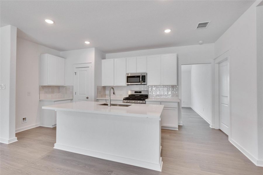 Spacious kitchen featuring an island with an integrated sink, white cabinetry, stainless steel appliances, light-toned countertops, and wood-finish flooring