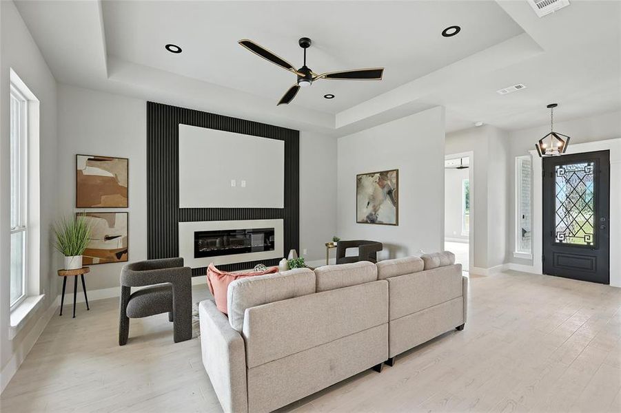 Living room featuring a raised ceiling, light wood-style floors, a glass covered fireplace, and recessed lighting