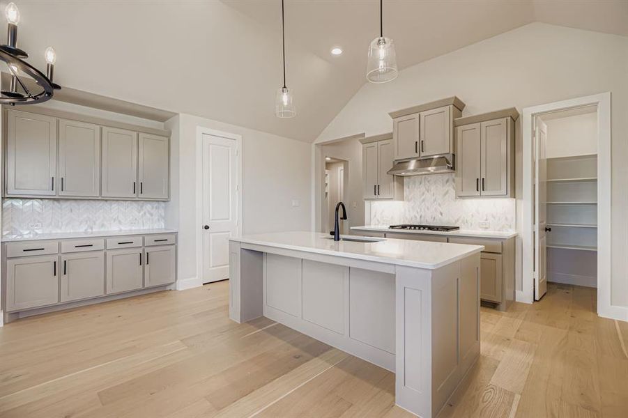 Kitchen featuring gray cabinets, vaulted ceiling, light wood-type flooring, tasteful backsplash, and light stone countertops