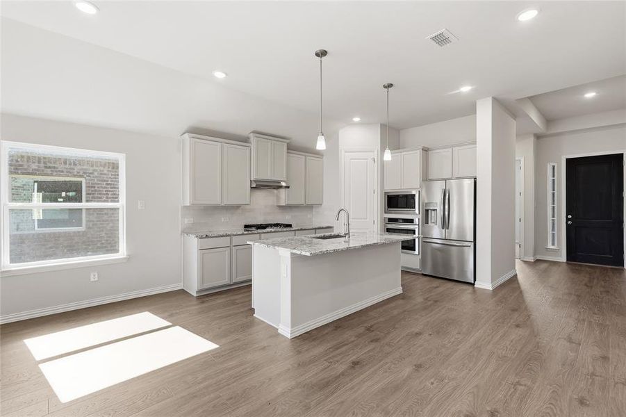 Kitchen featuring light stone counters, stainless steel appliances, hanging light fixtures, a center island with sink, and light wood-style floors Kitchen featuring light stone counters, stainless steel appliances, hanging light fixtures, a center island with sink, and light wood-style floors