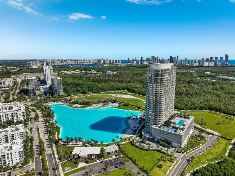 Aerial perspective of a high-rise residential tower featuring a multi-level podium, an attached swimming pool, landscaped grounds, and a central lagoon