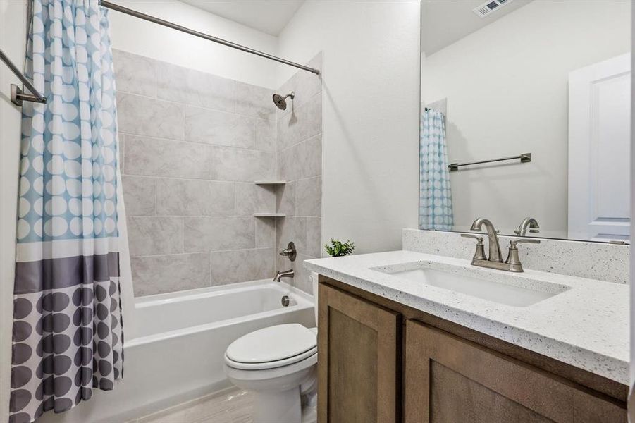 Full bathroom featuring a white tub, light-toned tiled shower surround with built-in shelving, a toilet, a vanity with wood-finish cabinetry, and a light-colored speckled countertop with an integrated sink