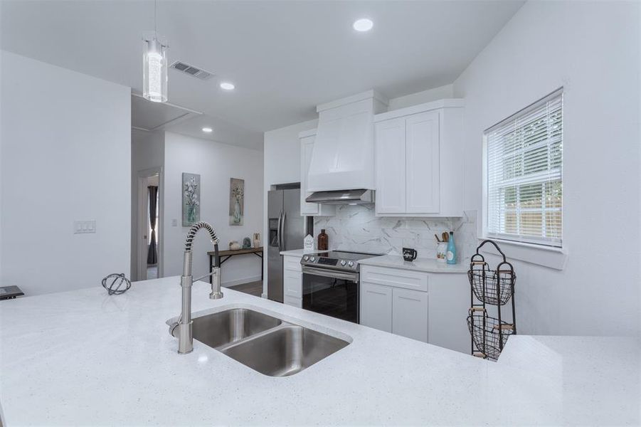 Kitchen featuring sink, white cabinetry, custom range hood, stainless steel appliances, and decorative light fixtures