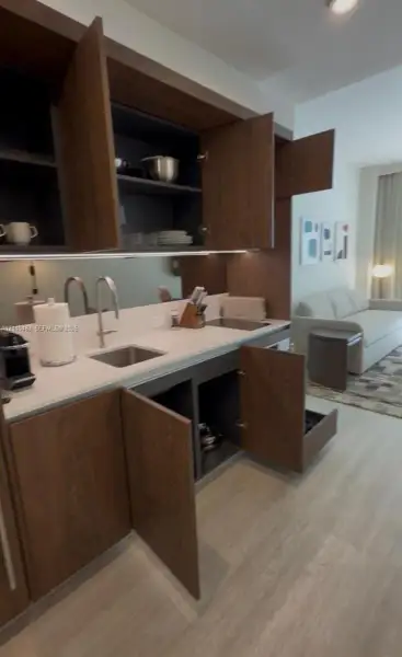 Custom kitchenette featuring dark wood-finish cabinetry, quartz countertops, a stainless steel undermount sink, integrated under-cabinet lighting, and a mirrored backsplash