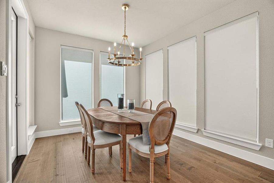 Dining space featuring wood-finish flooring, a gold-toned chandelier, multiple windows with roller shades, and light-toned wall paint Dining space featuring wood-finish flooring, a gold-toned chandelier, multiple windows with roller shades, and light-toned wall paint