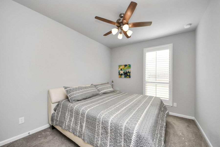 This bedroom features a cozy setup with a large bed, gray walls, and carpeted flooring. It includes a ceiling fan for comfort and a window with shutters allowing natural light.