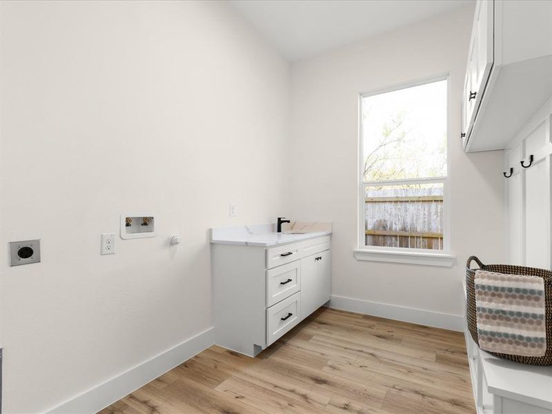 Laundry area featuring wood-finish flooring, a single window, white base cabinetry with a countertop and sink, and a built-in bench with upper cabinetry and hooks