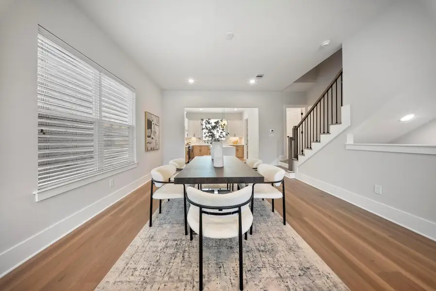 It is a bright, modern dining area with a sleek table and chairs, hardwood floors, and a view into a contemporary kitchen. The space is complemented by large windows, neutral walls, and a staircase with a stylish railing, adding to the open and inviting atmosphere.
