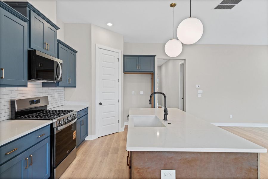 Kitchen featuring blue shaker cabinetry, stainless steel appliances, white subway tile backsplash, light-toned wood-finish flooring, and a central island with an integrated sink and matte black faucet Kitchen featuring blue shaker cabinetry, stainless steel appliances, white subway tile backsplash, light-toned wood-finish flooring, and a central island with an integrated sink and matte black faucet